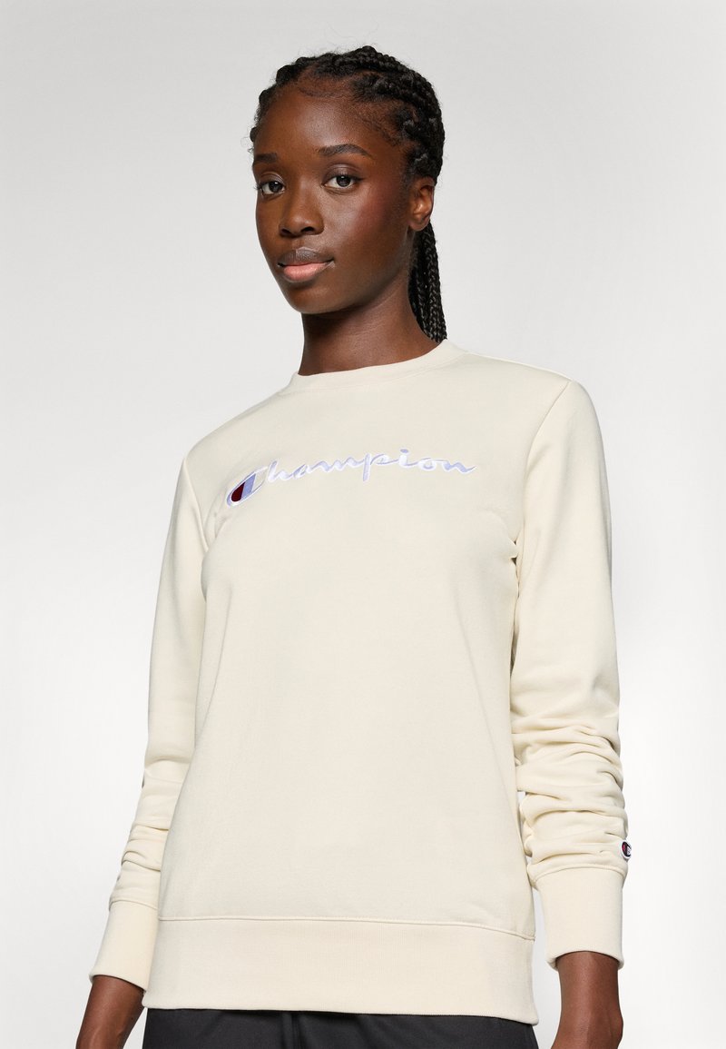 Cremfarbener Sweatshirt mit rundem Ausschnitt, gesticktem "Champion"-Logo in Lila auf der Brust und gerippten Bündchen und Saum.