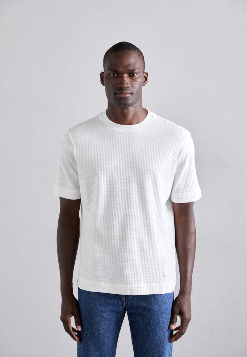 T-shirt blanc texturé à manches courtes avec un col rond, associé à un jean en denim bleu. Motif côtelé notable sur le tissu.