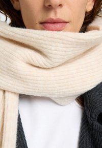 Ribbad gräddfärgad scarf gjord av mjuk ull, omsluten tätt runt halsen, med en lätt textur och subtil lyster.