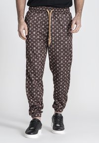 Pantalones jogger marrones con un diseño estampado y cintura con cordón, con un logotipo, tobillos ajustados y combinados con zapatillas negras.