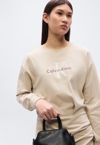 Μπεζ βαμβακερό φούτερ με ριμπ μανσέτες και στρογγυλή λαιμόκοψη, που φέρει ένα εμφανές λογότυπο σε λευκό και βουργουνδί. Το άτομο που το φορά κρατάει μια μαύρη δερμάτινη τσάντα.