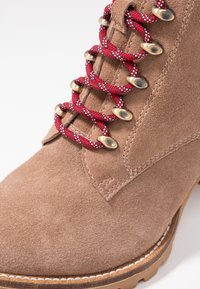 Brązowy zamszowy botek za kostkę z czerwono-białymi wzorzystymi sznurowadłami, metalowymi przelotkami i teksturowaną podeszwą. Charakteryzuje się widocznym przeszyciem i zaokrąglonym noskiem.