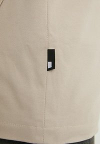 Beige tyg med diagonal twill-väv och en vertikal söm, med en liten svart och vit rektangulär etikett sydd i sömmen.