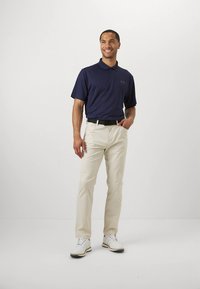 Tamsiai mėlynas trumpomis rankovėmis polo marškinėliai su faktūruotu raštu, kreminės kelnės, juodas diržas ir balti batai su akcentais. Stovintis vyriškas figūra.