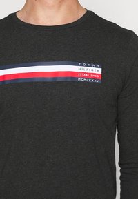 Schwarzes Langarm-T-Shirt aus weichem Stoff mit einem horizontalen gestreiften Logo in Rot, Weiß und Blau über der Brust.