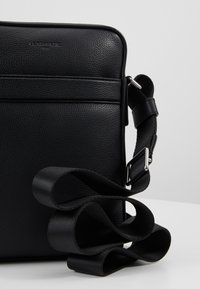 Borsa in pelle nera con finitura texture, dotata di una chiusura zip liscia e di una tracolla nera regolabile arrotolata accanto.