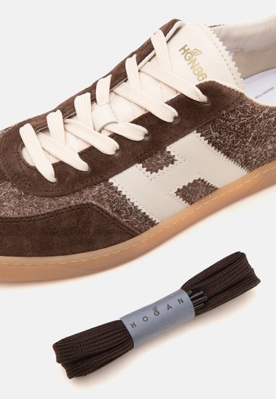 Baskets Hogan marron et beige avec des détails en suède texturé et cuir à côté d'une paire de lacets marron foncé enveloppés d'un ruban à l'effigie de la marque Hogan.