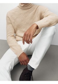 Beiger Strick-Rollkragenpullover mit Wellenmuster. Kombiniert mit weißen Hosen und dunklem Schuhwerk. Glatte Textur, figurbetontes Design.