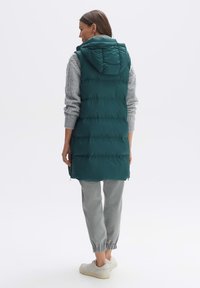 Tee-farbene gepolsterte Weste mit Kapuze, mit horizontalen Nähten, getragen über einem grauen Pullover und hellgrauen, schmal zulaufenden Hosen, kombiniert mit weißen Sneakern.