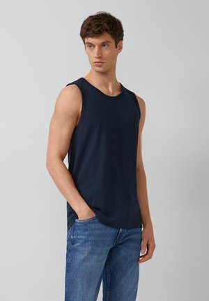 Junger Mann steht mit den Händen in den Taschen, trägt ein ärmelloses dunkelblaues Tanktop und blaue Jeans, schaut zur Seite vor einfarbigem Hintergrund.