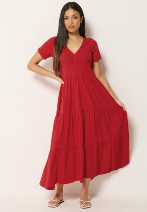 Femme debout en robe maxi rouge à volants avec manches courtes et décolleté en V, tenant l'ourlet de la robe, portant des sandales blanches sur fond uni.