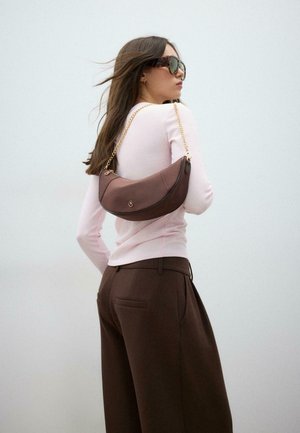 Femme aux cheveux longs portant des lunettes de soleil, un haut rose clair, un pantalon marron, portant un sac à main croissant marron avec une bandoulière chaîne dorée sur l'épaule.