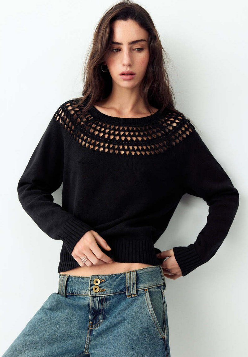 Femme portant un pull noir avec un empiècement à motifs et un jean bleu taille haute, debout, une main sur la hanche et regardant vers le bas.