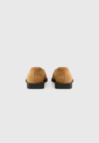 Καφέ μοκασίνια από σουέτ με χαμηλή μαύρη ελαστική σόλα, με λείες υφές και στρογγυλεμένη σιλουέτα, φαίνονται από πίσω.