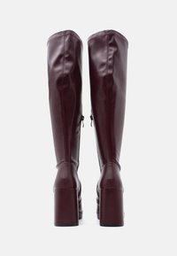 Μπορντό μπότες μέχρι το γόνατο από λείο δέρμα, με χοντρό τετράγωνο τακούνι και πλαϊνό φερμουάρ, με ορατές λεπτομέρειες ραφής.