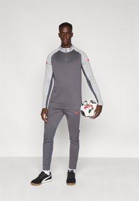 Γκρι αθλητικό μακρυμάνικο τοπ με μισό φερμουάρ και δίχρωμα μανίκια, συνδυασμένο με γκρι παντελόνια. Μαύρα αθλητικά παπούτσια. Κρατάει μια ποδοσφαιρική μπάλα με σχέδια.