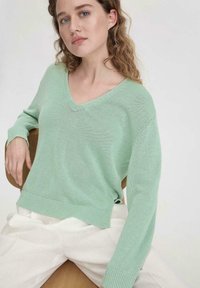 Hellgrüner V-Ausschnitt-Pullover mit gerippten Bündchen und Saum, aus strukturiertem Strickmaterial. Das Model sitzt auf einem Holzstuhl und trägt weiße Hosen.