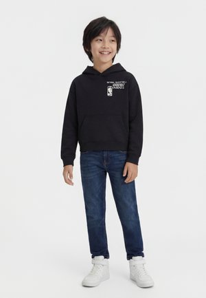 Fant deček, ki se smeji, oblečen v črno kapuco NBA, modre kavbojke in bele superge z visokim zgornjim delom, stoji pred belim ozadjem.