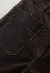 Ruskeat corduroy-housut, joissa on takatasku suorakaiteen muotoinen, näkyvillä ompeleilla ja teksturoidulla pinnalla.