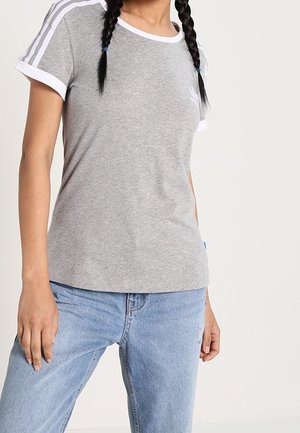 Osoba ubrana w szarą koszulkę z krótkim rękawem z białymi paskami na ramionach i jasnoniebieskie dżinsy, stojąca na jednolitym tle.