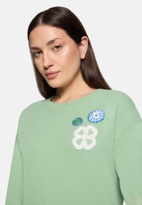 Jasnozielony sweter z miękkiego materiału z czterema wyraźnymi haftowanymi naszywkami w niebieskie i białe wzory geometryczne.