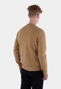 Ribbet, beige genser med rund hals, lange ermer, og en tettsittende kant. Har subtil oransje branding nederst til venstre.