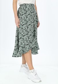 Jupe florale noire avec un ourlet évasé, présentant des motifs de fleurs vert clair. Portée avec des baskets blanches sur un fond uni.