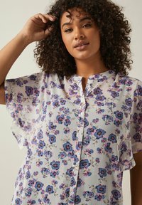 Leichte, transparente Bluse in Weiß mit floralem Muster in Blau und Pink. Verfügt über eine durchgehende Knopfleiste und kurze, schwingende Ärmel.