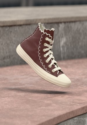 Wysoki, bordowy sneaker z białym falistym przeszyciem i sznurówkami, biała gumowa podeszwa i ochronny czubek, zawieszony nad kamienną powierzchnią.