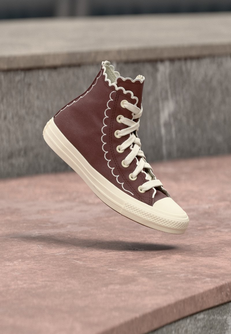 Μαύρο-κόκκινο αθλητικό παπούτσι ψηλού τύπου με λευκό κυματιστό ράψιμο και κορδόνια, λευκή λαστιχένια σόλα και προστασία μύτης, κρεμασμένο πάνω από μια πέτρινη επιφάνεια.