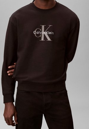 Homme portant un sweat-shirt Calvin Klein noir à manches longues avec un grand logo et texte "cK" sur la poitrine, pantalon noir, debout devant un fond gris.