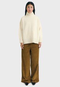 Young woman wearing a cream turtleneck sweater, brown wide-leg corduroy pants, and black pointed shoes standing against a plain background.