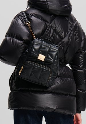 Quiltad svart läderryggsäck med guldfärgade beslag, med ett huvudfack och en framficka med dragkedja, bärs över en puffjacka.