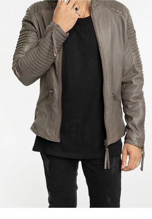 Homme portant une veste en cuir matelassée grise sur une chemise noire et un jean noir déchiré, avec des bagues à la main gauche près du visage.