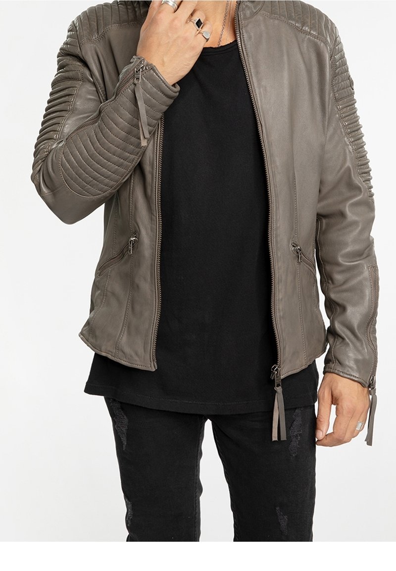 Homme portant une veste en cuir matelassée grise sur une chemise noire et un jean noir déchiré, avec des bagues à la main gauche près du visage.