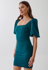 Teal short dress with textured fabric, square neckline, and puff sleeves. Fitted design with a smooth finish. Gold earrings add contrast.