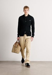 Fekete kötött kardigán, bézs nadrág, barna táska. A modellen fekete cipők vannak zöld csíkokkal. Egyszerű, semleges háttér.