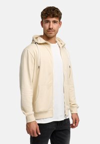 Lichtbeige hoodie met rits, gemaakt van zacht materiaal met een kangoeroezak, verstelbare capuchon met trekkoord en geribde boorden, gedragen over een wit shirt.