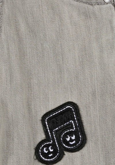 Écusson noir brodé en forme de note musicale avec deux visages souriants cousus sur un fond de tissu gris.