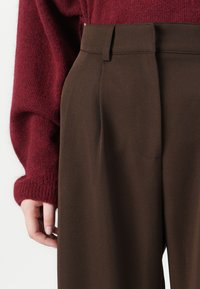 Braune, maßgeschneiderte Hose mit glatter Textur, mit einem taillierten Bund, Falten und seitlichen Taschen. Ein weicher roter Strickpullover ist teilweise sichtbar.