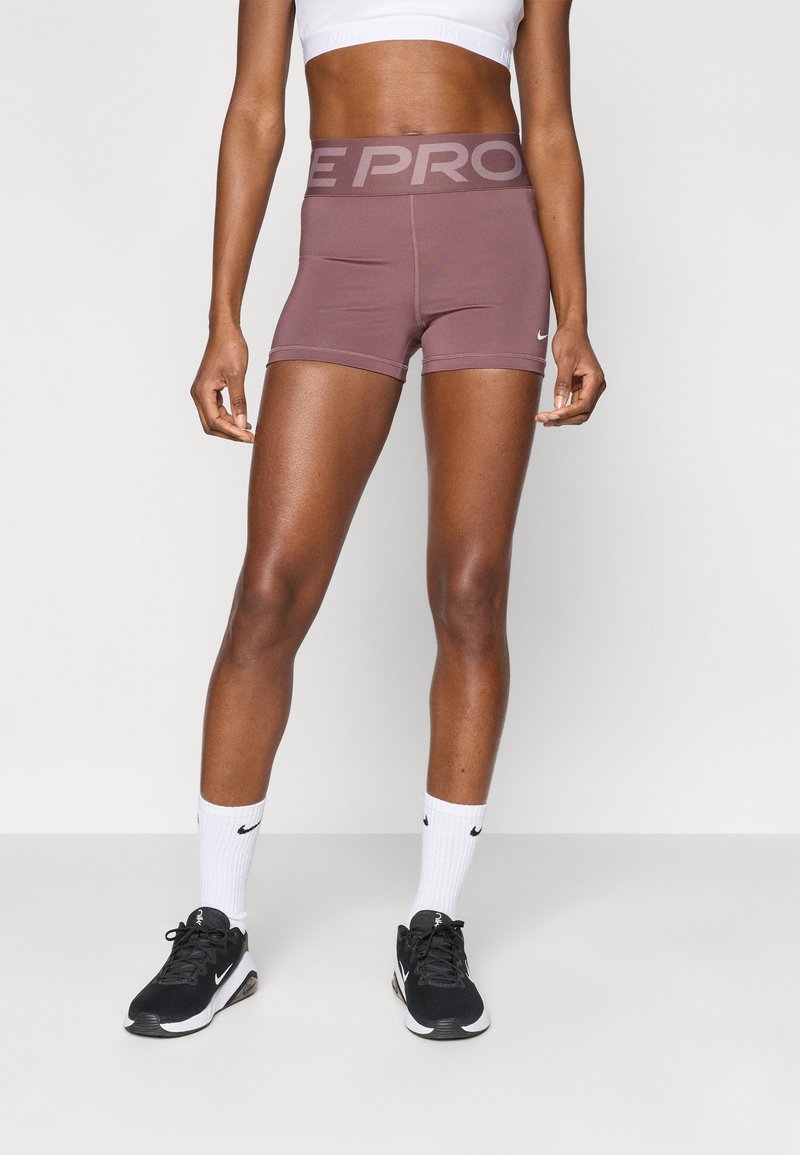 Σορτς Nike Pro, κοντό μήκος, μονόχρωμο μωβ, ελαστική μέση με το κείμενο "PRO", λεία, ελαστική ύφανση, συνδυασμένα με μαύρα αθλητικά παπούτσια.