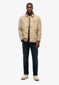 Beige Jacke mit Knopfverschluss und vorderen Taschen, über einem karierten Hemd getragen; kombiniert mit dunklen Jeans und hellen Sneakers. Einfaches Design, strukturiertes Material.