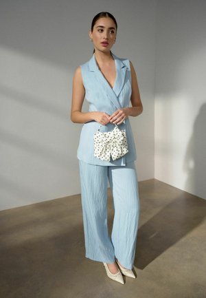 Frau in hellblauem ärmellosem Blazer und Hose, die eine weiße Handtasche mit Polka-Dot-Schleife hält, steht drinnen im weichen natürlichen Licht.