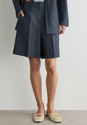 Kvinde iført marineblå skræddersyede knælange shorts, matchende jakke og cremefarvede balletsko med sløjfer, stående mod en ensfarvet væg.