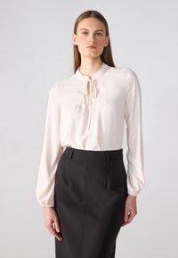 Blusa rosa claro con mangas largas y abullonadas y un collar tipo mandarín, que presenta delicados pliegues y un cierre de lazo en el escote, combinada con una falda negra.
