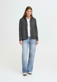 Grijs textuurgevoerd jack met rits en capuchon, gecombineerd met een witte top en lichtblauwe jeans. Het jack heeft zwarte afwerking en een ontspannen pasvorm.