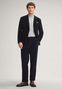 Marineblauwe corduroy blazer met notch-lapels, gecombineerd met bijpassende broeken. Grijze getailleerde coltrui eronder. Bruine leren laarzen.