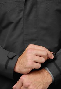 Černá bunda s texturovanou látkou, nastavitelné manžety na suchý zip a hladká vnitřní podšívka. Detail rukou zapínajících manžetu.