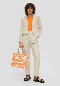 Beige cropped blazer over een oranje top, gecombineerd met beige cargo pants. Een grote oranje en witte gestreepte tote bag met een grijze handgreep maakt de look compleet.