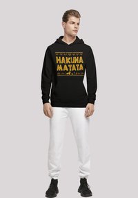 Svart huvtröja med "HAKUNA MATATA" i gul text, med tribalmönster. Matcher med vita sweatpants och svarta sneakers.